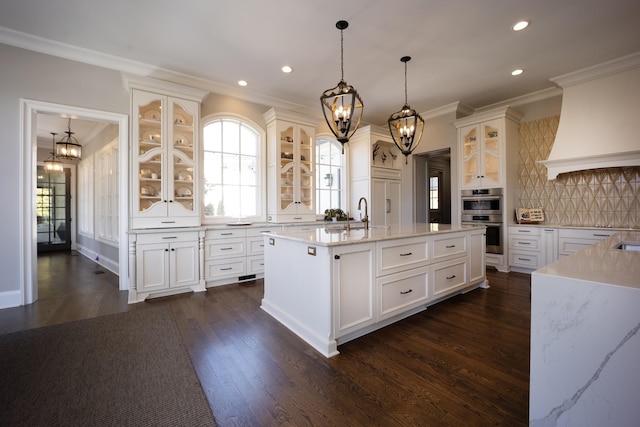 kitchen featuring custom exhaust hood, tasteful backsplash, ornamental molding, stainless steel double oven, and a kitchen island with sink