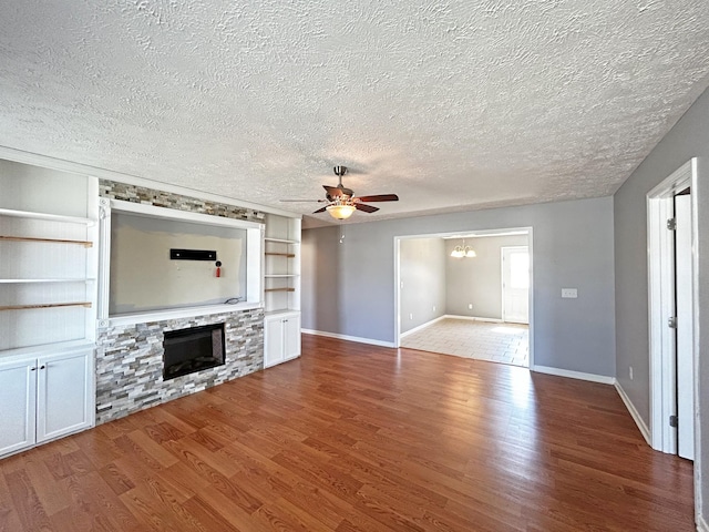 unfurnished living room with built in features, a stone fireplace, ceiling fan with notable chandelier, wood finished floors, and a textured ceiling