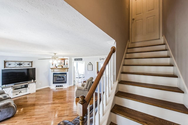 stairs featuring a textured ceiling, wood finished floors, a ceiling fan, baseboards, and a glass covered fireplace