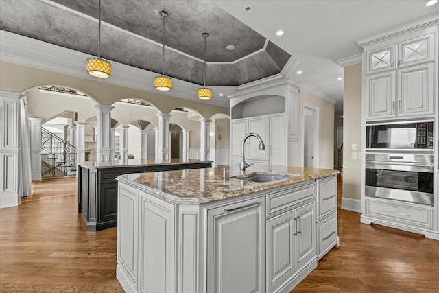 kitchen with decorative columns, stainless steel oven, a sink, an island with sink, and black microwave