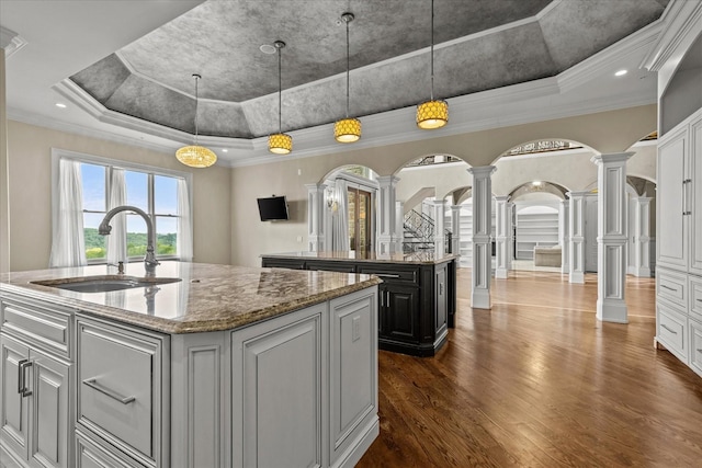 kitchen with arched walkways, a tray ceiling, a kitchen island with sink, a sink, and ornate columns