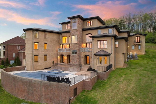 back of property at dusk featuring a lawn, a patio, a balcony, fence, and stucco siding