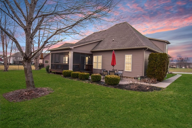 back of house at dusk with a sunroom, roof with shingles, a lawn, and a patio