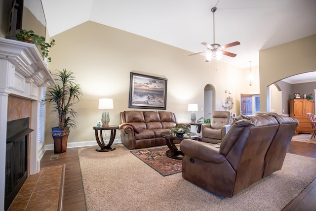 living area with arched walkways, dark wood finished floors, a tiled fireplace, ceiling fan, and high vaulted ceiling