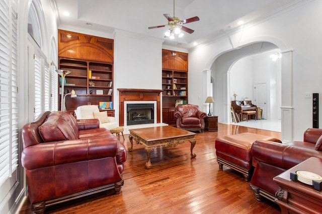 living area featuring built in shelves, arched walkways, crown molding, a fireplace, and wood-type flooring