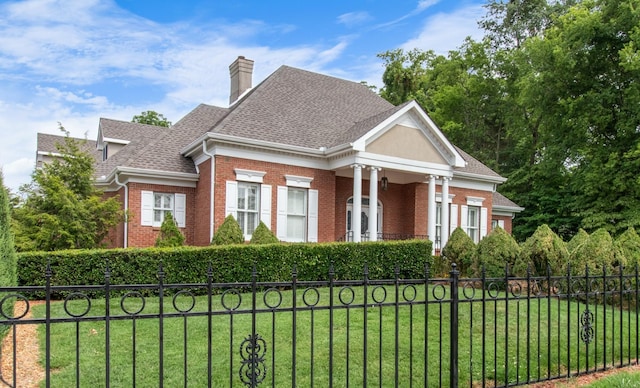 neoclassical / greek revival house with a fenced front yard, brick siding, roof with shingles, a chimney, and a front yard