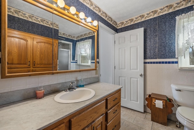 bathroom with toilet, tile walls, vanity, a shower stall, and wallpapered walls