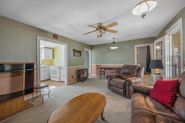 living room with a ceiling fan, a wood stove, light carpet, baseboards, and wallpapered walls