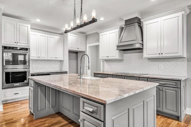 kitchen with custom exhaust hood, gray cabinets, stainless steel double oven, a kitchen island with sink, and a sink