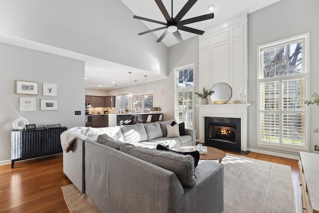 living area featuring a large fireplace, baseboards, a ceiling fan, dark wood-style floors, and high vaulted ceiling