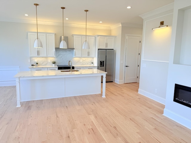 kitchen with a sink, light wood finished floors, stainless steel fridge, a center island with sink, and tasteful backsplash