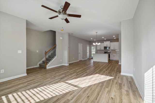 unfurnished living room with recessed lighting, ceiling fan with notable chandelier, baseboards, stairs, and light wood-type flooring