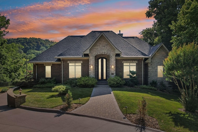 french country home with brick siding, a shingled roof, stone siding, and a yard