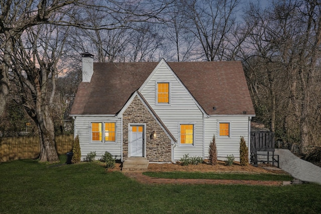 cape cod home featuring a chimney, a shingled roof, a front yard, fence, and stone siding