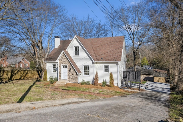 cape cod home featuring a chimney, a shingled roof, a front yard, fence, and stone siding