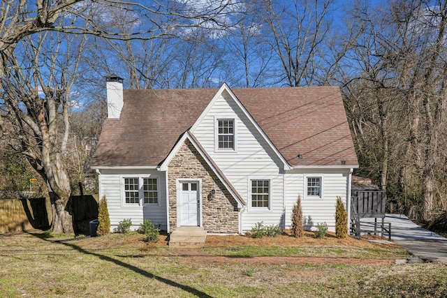 cape cod-style house featuring fence, stone siding, roof with shingles, a front lawn, and a chimney