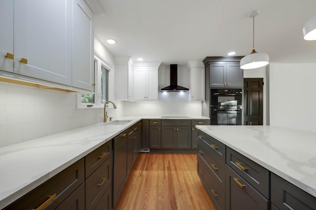 kitchen with dobule oven black, a sink, light wood-style floors, light stone countertops, and wall chimney exhaust hood