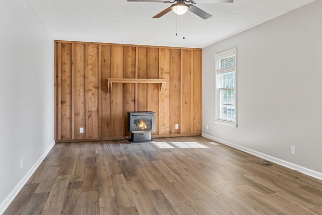 unfurnished living room featuring a ceiling fan, a wood stove, wooden walls, wood finished floors, and baseboards