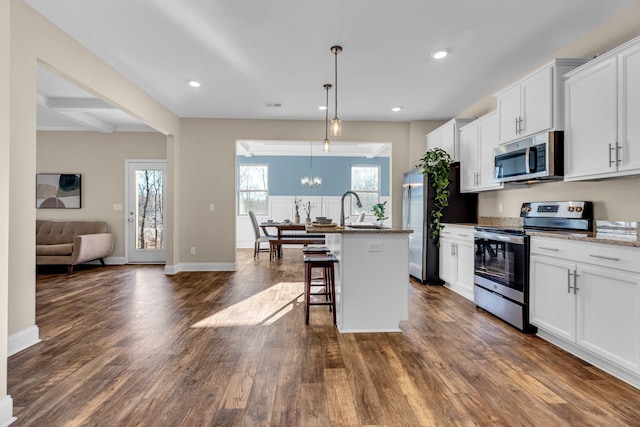 kitchen with dark wood finished floors, appliances with stainless steel finishes, a kitchen breakfast bar, a kitchen island with sink, and a notable chandelier