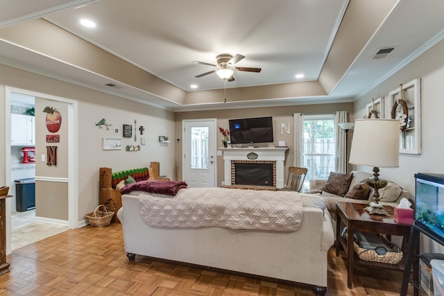 living area featuring visible vents, ceiling fan, a tray ceiling, crown molding, and a brick fireplace