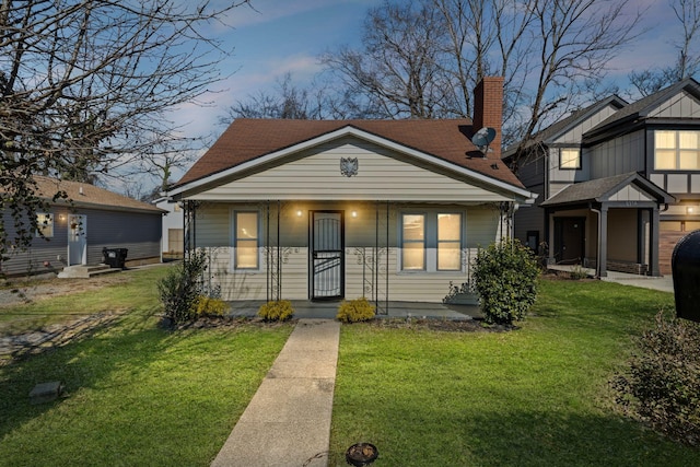 bungalow-style house featuring covered porch, a shingled roof, a chimney, and a lawn