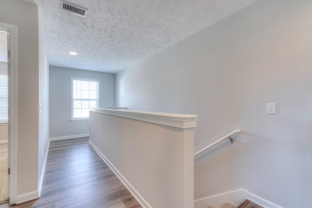 hall featuring baseboards, visible vents, wood finished floors, a textured ceiling, and an upstairs landing