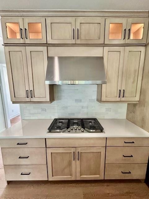 kitchen with tasteful backsplash, wall chimney exhaust hood, glass insert cabinets, light countertops, and stainless steel gas stovetop