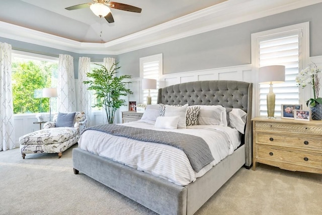 carpeted bedroom featuring a wainscoted wall, crown molding, a raised ceiling, a decorative wall, and a ceiling fan