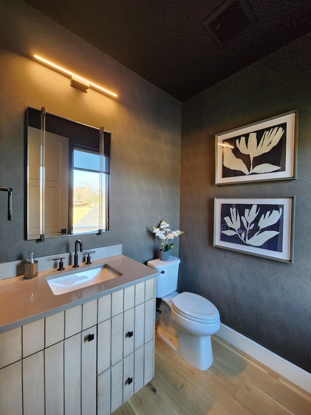 half bathroom with toilet, wood finished floors, vanity, visible vents, and wallpapered walls