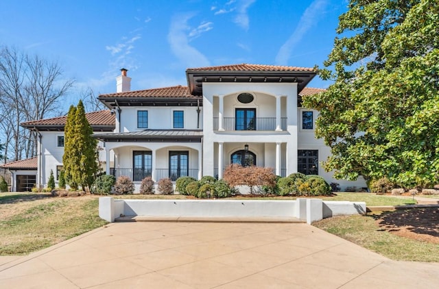 mediterranean / spanish house featuring a balcony, stucco siding, a chimney, metal roof, and a front yard