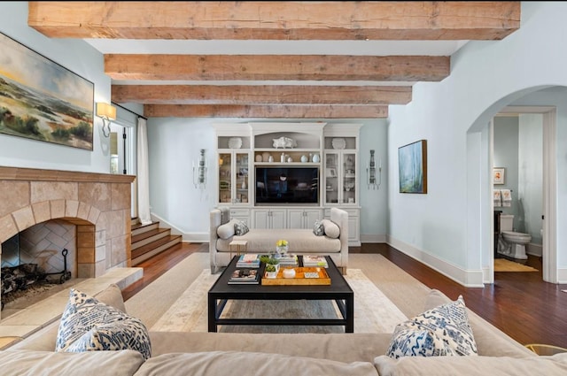 living room with arched walkways, wood finished floors, a tiled fireplace, and beamed ceiling