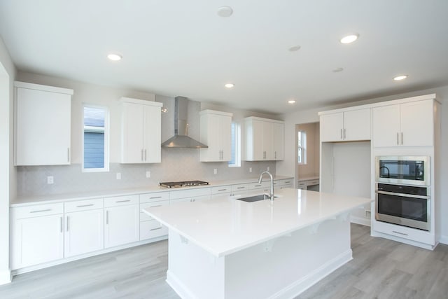 kitchen with appliances with stainless steel finishes, white cabinets, a sink, wall chimney range hood, and an island with sink