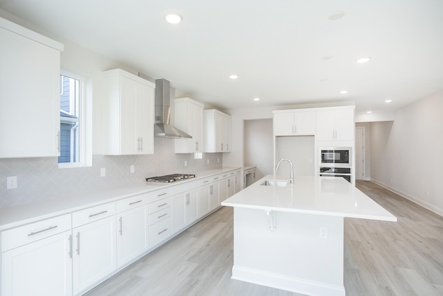 kitchen with stainless steel appliances, backsplash, light wood-style flooring, a sink, and wall chimney exhaust hood