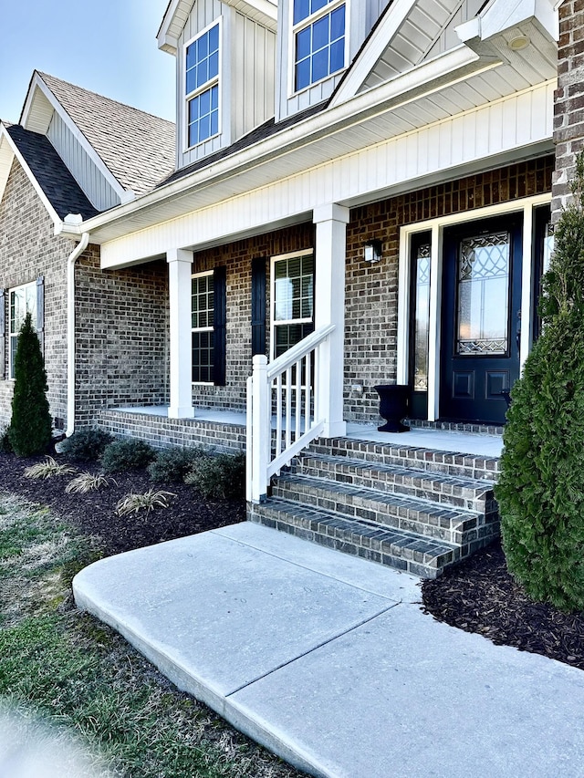 doorway to property featuring covered porch, a shingled roof, board and batten siding, and brick siding