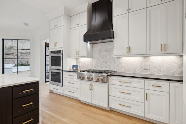 kitchen with custom exhaust hood, stainless steel appliances, light wood-type flooring, white cabinetry, and backsplash