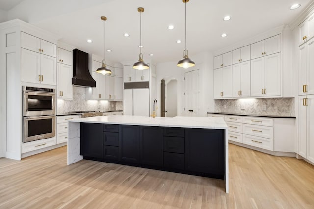 kitchen featuring arched walkways, stainless steel appliances, a spacious island, custom exhaust hood, and light wood finished floors