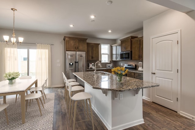 kitchen with a breakfast bar area, dark wood-style flooring, a kitchen island with sink, stainless steel appliances, and a notable chandelier