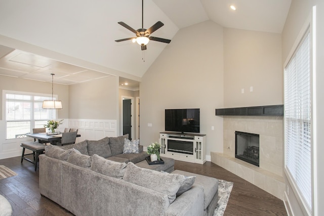 living room with high vaulted ceiling, a tile fireplace, dark wood-type flooring, a ceiling fan, and wainscoting