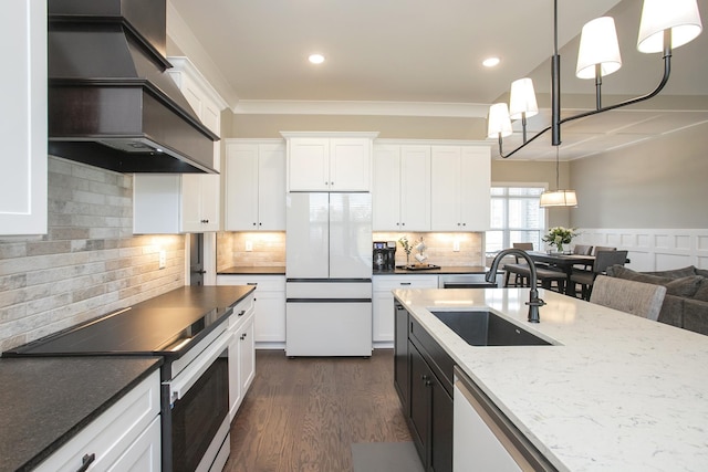 kitchen with white appliances, white cabinets, custom range hood, crown molding, and a sink