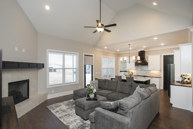 living room featuring baseboards, a tile fireplace, dark wood-type flooring, high vaulted ceiling, and ceiling fan with notable chandelier