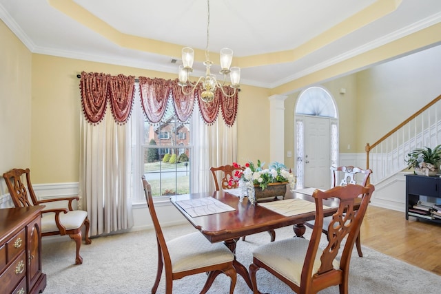 dining area with a wainscoted wall, a notable chandelier, a raised ceiling, ornate columns, and stairs