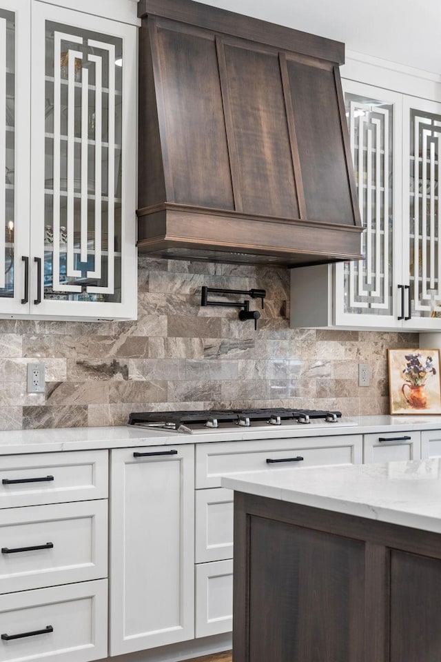 kitchen featuring stainless steel gas cooktop, premium range hood, white cabinets, decorative backsplash, and glass insert cabinets