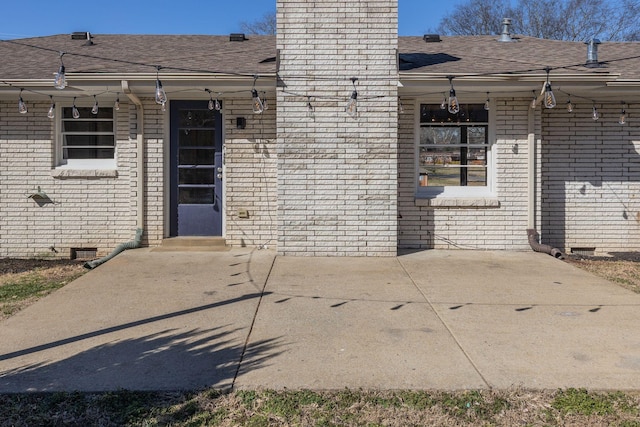 view of exterior entry featuring crawl space, a shingled roof, a chimney, and brick siding