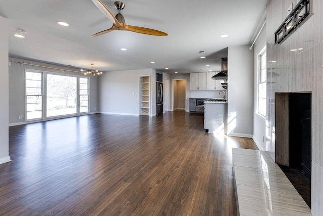 unfurnished living room featuring baseboards, dark wood-style flooring, ceiling fan with notable chandelier, a sink, and recessed lighting