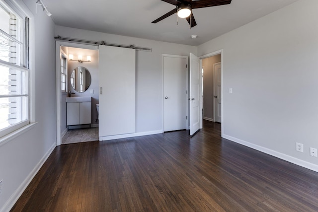 unfurnished bedroom featuring a barn door, connected bathroom, baseboards, and dark wood-type flooring