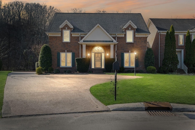 view of front facade featuring driveway, brick siding, roof with shingles, and a yard