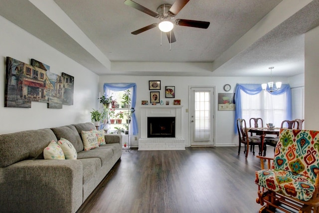 living area featuring dark wood-style floors, ceiling fan with notable chandelier, a brick fireplace, and a raised ceiling