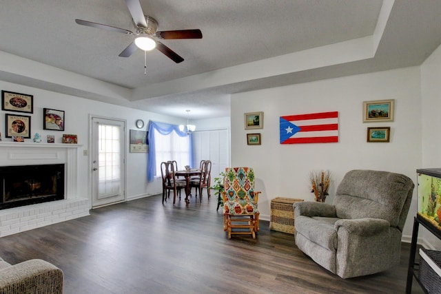 living area featuring a tray ceiling, dark wood-type flooring, a brick fireplace, baseboards, and ceiling fan with notable chandelier