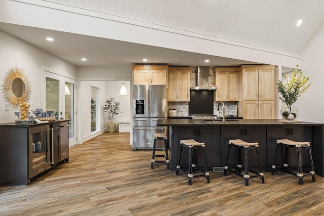 kitchen with stainless steel fridge, dark countertops, wall chimney exhaust hood, wood finished floors, and a kitchen bar