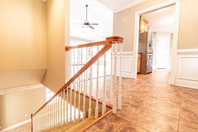 stairs with ceiling fan, ornamental molding, a wainscoted wall, and a decorative wall